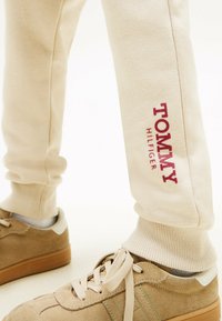 Beige Sweatpants mit gerippten Bündchen, versehen mit dem roten "TOMMY HILFIGER" Logo am linken Bein, kombiniert mit hellbraunen Wildledersneakern.