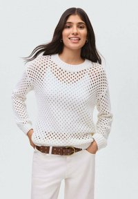 Weißer Strickpullover mit Lochmuster-Design, geripptem Kragen und Bündchen. Getragen mit hellen Hosen und einem braunen gepunkteten Gürtel.