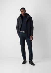 Marineblauwe waterdichte jas met een capuchon, gestructureerde stof en voorzakken, gecombineerd met donkere broek en zwarte veterschoenen. Minimalistisch ontwerp.