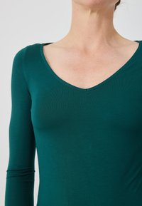 Dugi rukav majica u tirkiznoj zelenoj boji, izrađena od glatkog materijala s dubokim V-izrezom i prilagođenim oblikom, koja pokazuje blagu teksturu na površini.