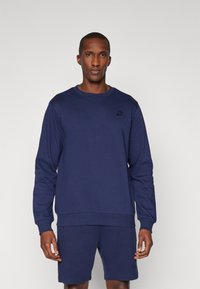 Marineblaues Langarmshirt mit Rundhalsausschnitt, gerippten Bündchen und Saum, mit einem dezenten Logodesign auf der Brust. Kombiniert mit passenden Shorts.
