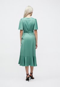 Πράσινο σατέν φόρεμα με κοντά φουσκωτά μανίκια, με καθορισμένη ζώνη στη μέση και φαρδύ στρίφωμα. Λείο υλικό; φορεμένο με μαύρες πλατφόρμες.