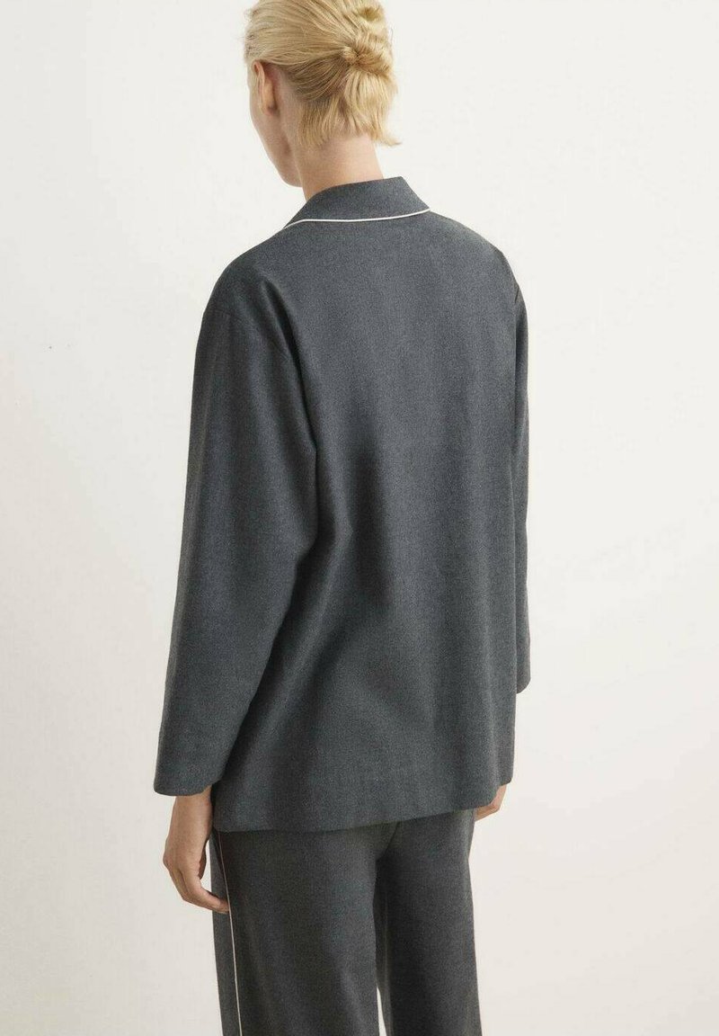 Persona con cabello rubio corto, vistiendo una camiseta de manga larga suelta de color gris oscuro y pantalones a juego, de espaldas contra un fondo claro liso.