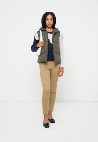 Gepolsterte olivgrüne Weste über einem grauen und marineblauen Pullover, kombiniert mit Khakihosen und schwarzen Ballerinas. Hält eine weiße Handtasche.