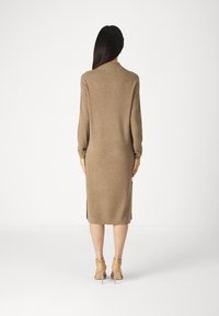 Abito in maglia marrone con maniche lunghe, spacchi laterali e scollatura rotonda, abbinato a scarpe trasparenti con tacco alto. Texture liscia.