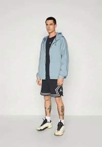 Ljust blå zip-up jacka med huva över en svart T-shirt, kombinerad med svarta shorts med vita detaljer och sportiga skor.