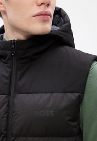 Melns kapuci bezr集 vestē ar quilted tekstūru un diskretu "BOSS" logo krūtīs. Ar rāvējslēdzēja aizdari un gludu auduma apdari.
