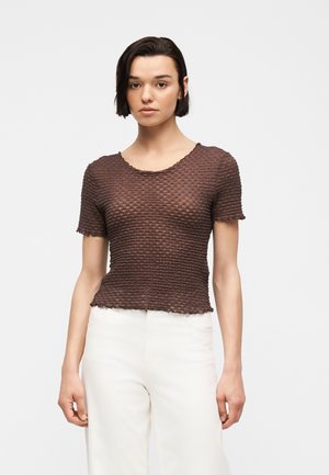 Jeune femme aux cheveux courts et foncés, portant un haut marron texturé à manches courtes et un pantalon blanc taille haute, se tenant devant un fond uni.