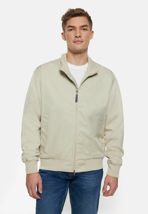 Beige letvægtsjakke med lynlås foran, opretstående krave, elastiske manchetter og sidelommer, båret over en hvid T-shirt. Glat stof tekstur.