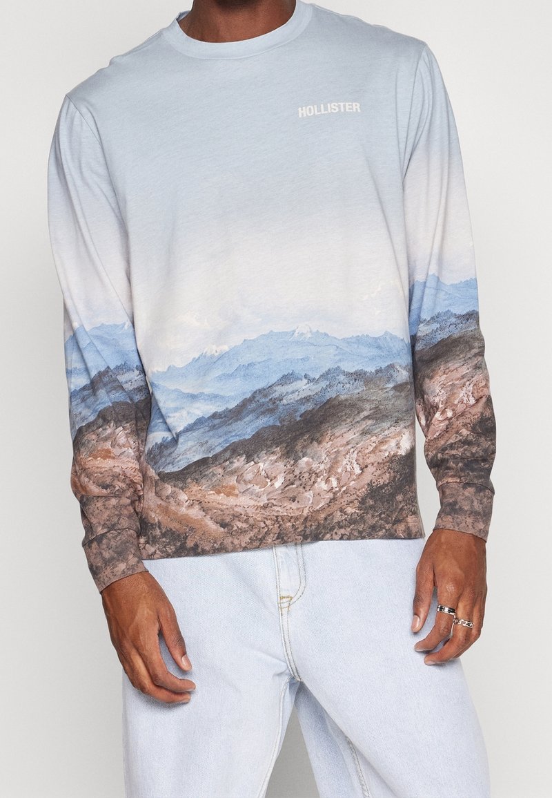 Langarmhemd in Hellblau mit einem Berglandschaftsprint in Blau- und Brauntönen; „HOLLISTER“-Logo auf der Brust.