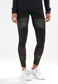 Czarne legginsy z zielonymi paskami w tygrysie prążki, siateczkowymi akcentami i dopasowanym krojem. Noszone z białymi butami sportowymi.