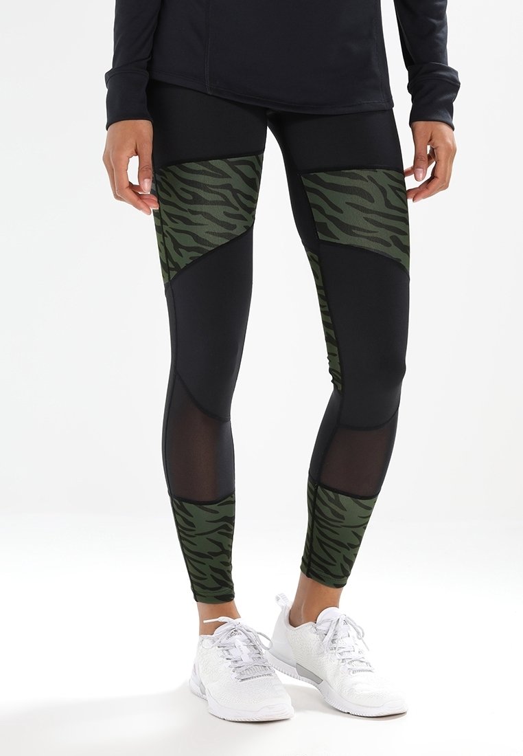 Czarne legginsy z zielonymi paskami w tygrysie prążki, siateczkowymi akcentami i dopasowanym krojem. Noszone z białymi butami sportowymi.