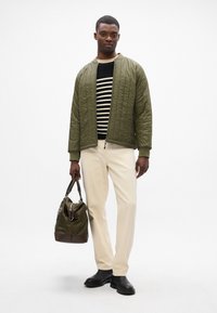 Giacca bomber trapuntata verde oliva su un maglione a righe, abbinata a pantaloni color crema, scarpe nere e una borsa verde con dettagli marroni.