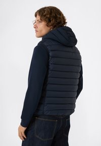 Młody mężczyzna z kręconymi włosami, ubrany w granatową pikowaną kamizelkę z kapturem na długim rękawie i ciemnych dżinsach, zwrócony w lewo na białym tle.