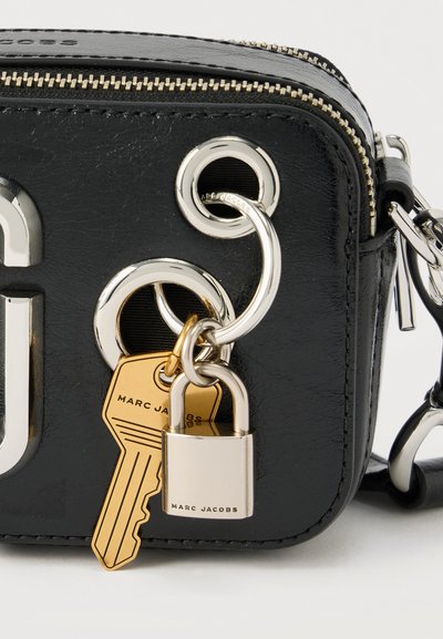 Γωνία μαύρης δερμάτινης τσάντας με ασημένιο φερμουάρ, ασημένιες κρίκους, ένα χρυσό φυλαχτό σε σχήμα κλειδιού και ένα ασημένιο φυλαχτό σε σχήμα λουκέτου με χαραγμένο το όνομα "Marc Jacobs."