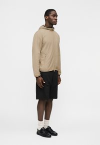 Άνδρας στέκεται φορώντας μπεζ μπουφάν με κουκούλα, μαύρο σορτς, λευκές κάλτσες και μαύρα αθλητικά παπούτσια σε απλό λευκό φόντο.