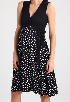 Femme portant une robe noire sans manches à pois blancs, cintrée à la taille avec une ceinture nouée, jupe longueur genou.