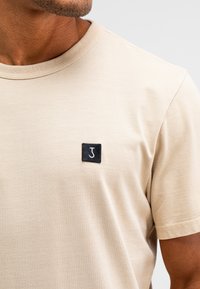 Beige T-shirt met korte mouwen, gemaakt van zachte stof, met een zwart vierkantlabel met een witte gestileerde "J"-logo op de linkerkant van de borst.