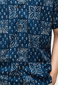 Μπλε κοντομάνικο πουκάμισο με κουμπιά, διακοσμημένο με λευκά αφηρημένα φύλλα και ρίγες ζέβρας, φορεμένο από άτομο με το ένα χέρι στην τσέπη.