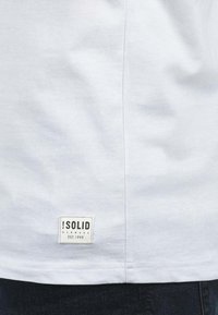 Hvid bomulds t-shirt med en glat tekstur, der har et rektangulært mærke ved kanten, som skriver "!SOLID, DANMARK, EST. 1999."
