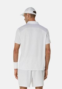 Witte sportieve polo met mesh schouderdetails, gecombineerd met bijpassende shorts en een witte pet, vergezeld van een polsband om de linkerarm.