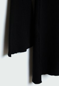 Pull noir en maille côtelée avec un col montant et un ourlet asymétrique. La texture est douce avec des côtes visibles et des détails subtils sur les bords.