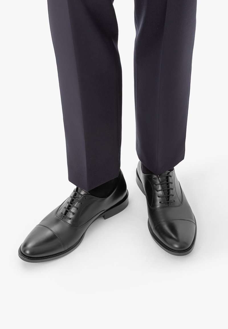 Chaussures habillées en cuir noir avec lacets portées avec un pantalon formel foncé sur fond blanc.