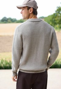 Grå ribbet sweater med krave, der har lange ærmer og en afslappet pasform, parret med mørke bukser, sat mod en udendørs baggrund.