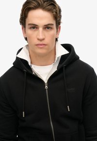 Schwarze Kapuzenjacke mit Reißverschluss aus weichem Stoff, ausgestattet mit einer Kordelzugkapuze, vorderen Taschen und einem darunter sichtbaren weißen Hemd mit Kragen.