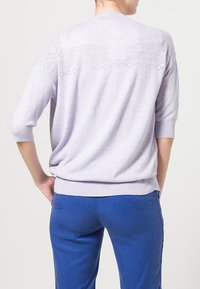 Lavendelstickad tröja med korta ärmar och öppna geometriska mönster på ryggen. Kombinerad med blå byxor. Mjuk textur.