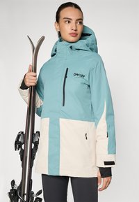 Ski jacket in teal and cream, with a hood, zippered pocket, and velcro cuffs. Smooth material with sleek design, featuring a branding logo.