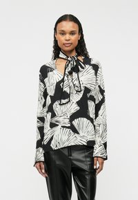 Blusa a maniche lunghe nera con motivi a foglia bianchi. Presenta un collo a V e un nastro nero annodato al collo. Tessuto morbido e leggero.