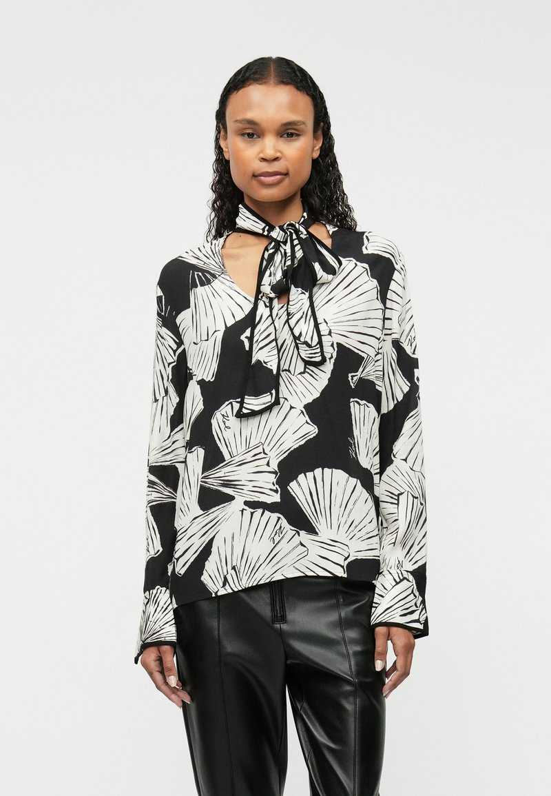 Blusa a maniche lunghe nera con motivi a foglia bianchi. Presenta un collo a V e un nastro nero annodato al collo. Tessuto morbido e leggero.