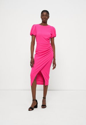 Kobieta ubrana w jasnoróżową sukienkę midi z krótkimi bufkami i kopertowym dołem, w parze z czarnymi sandałami na słupku z paskiem wokół kostki.