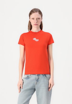 T-shirt rouge à manches courtes avec un col rond et un petit logo blanc au centre. Le tissu est lisse et extensible ; à associer avec un jean clair.