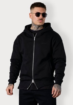 Black hoodie with a zippered front, ribbed cuffs and hem, minimalist design, featuring a subtle logo and layered with a crew neck shirt.