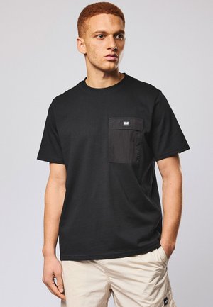 Junger Mann mit kurzen lockigen Haaren, der ein schwarzes T-Shirt mit Brusttasche und beiger Shorts vor einem schlichten hellen Hintergrund trägt.