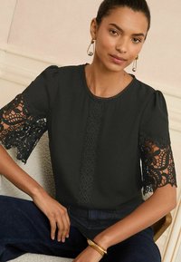 Blusa nera con maniche corte a palloncino, caratterizzata da dettagli di pizzo e ricami intricati lungo le maniche e al centro. Finitura in tessuto testurizzato.