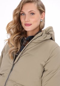 Vrouw met golvend blond haar, die zilveren oorbellen draagt, een beige gewatteerde jas met capuchon en een bruin-blauw geruite shirt eronder, die iets glimlacht.