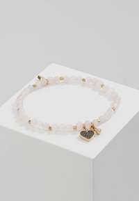 Bracelet en perles de quartz rose avec des accents dorés. Comprend un charm en forme de cœur noir et un petit bijou transparent, posé sur un piédestal d'exposition blanc.