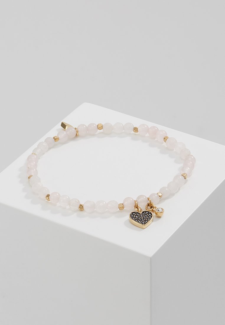 Bracelet en perles de quartz rose avec des accents dorés. Comprend un charm en forme de cœur noir et un petit bijou transparent, posé sur un piédestal d'exposition blanc.