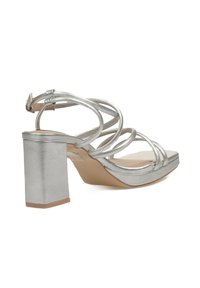 Sandalias de tacón plateado con tiras entrelazadas, tacón cuadrado, textura suave y un diseño minimalista. Suela plana con acentos en marrón claro.