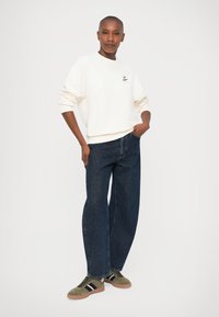 Felpa crema, vestibilità rilassata con polsini a costine. Jeans in denim scuro, modello loose straight-leg. Sneakers verde oliva con strisce nere e suola in gomma.