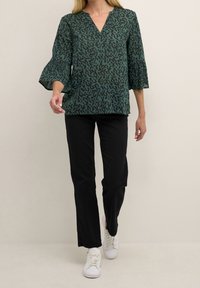 Blouse à motifs verts avec col en V et manches évasées, associée à un pantalon noir et des baskets blanches. Tissu lisse et coupe décontractée.