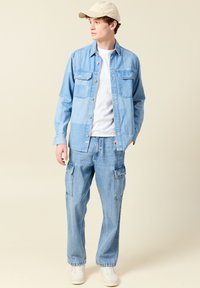 Vaaleansininen farkkupaita, jossa on kaksi rintataskua, valkoisen t-paidan päällä. Rennot vaaleansiniset cargo-housut ja beige lippis.