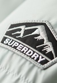 Logotipo bordado en blanco y negro sobre una suave tela gris claro, que presenta formas angulares y la palabra "SUPERDRY" en texto claro y en negrita.