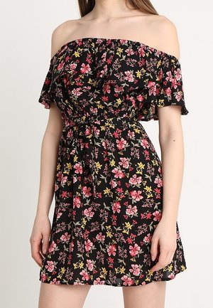 Nainen pukeutuneena mustaan olkapäät paljastavaan mekkoon, jossa on vaaleanpunaisia, punaisia ja keltaisia kukkakuoseja ja röyhelöinen pääntie.