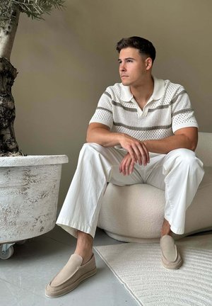 Jonge man in witte broek en gestreept gebreid shirt die op een lichte stoel naast een grote plant in een pot zit, nadenkend naar de zijkant kijkend.