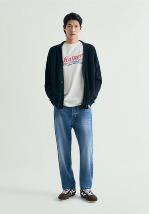 Bărbat tânăr în picioare, purtând un cardigan negru, un tricou alb cu grafică, blugi albaștri și adidași maro pe un fundal simplu și deschis.