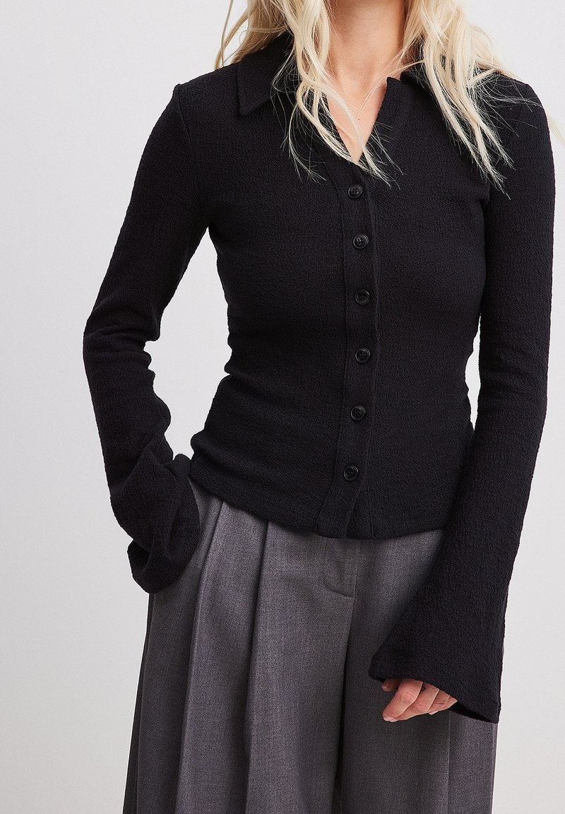 Zwarte blouse met lange mouwen en knopen, gemaakt van een gestructureerde stof, met een kraag en uitlopende manchetten, gecombineerd met grijze wijde broekspijpen.